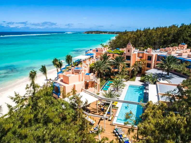 Aerial view of SALT of Palmar in Mauritius, with coral-pink buildings, a pool, palm trees, and turquoise beachside waters.