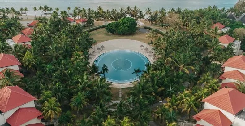 Aerial view of Tamassa Mauritius with a round pool, palm trees, and red-roofed villas near the beach.