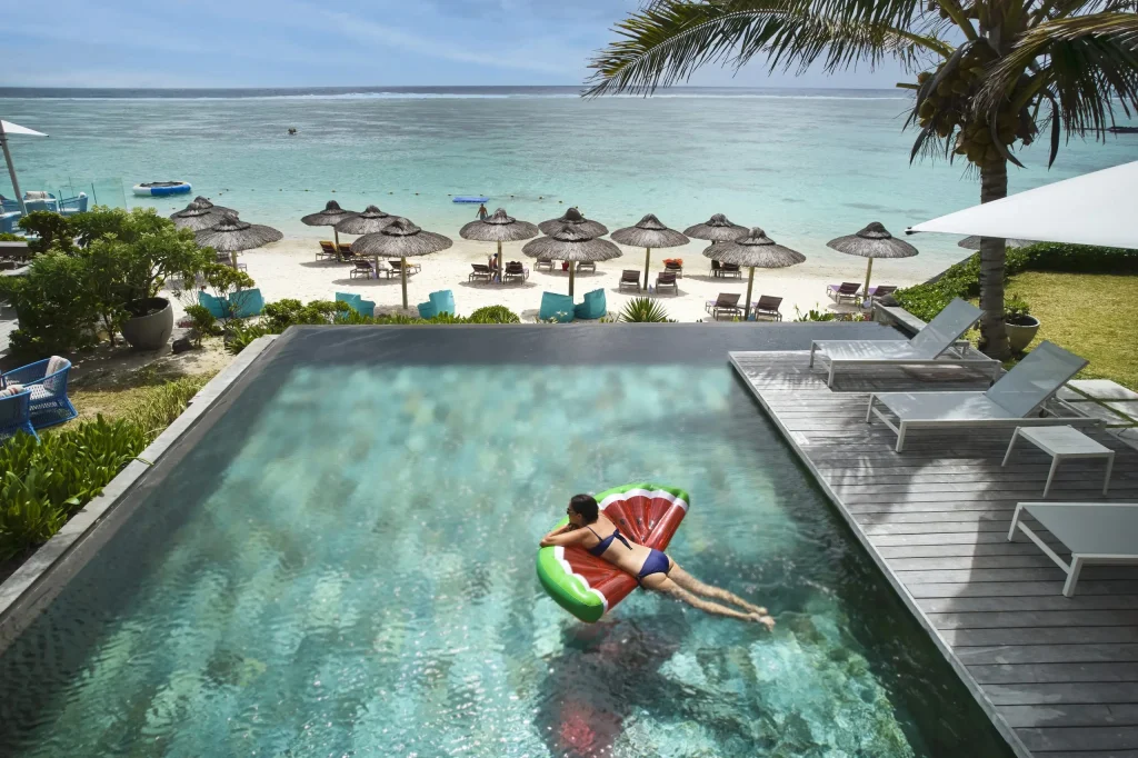 A woman floats on a watermelon pool float in an infinity pool overlooking the turquoise ocean at C Mauritius resort