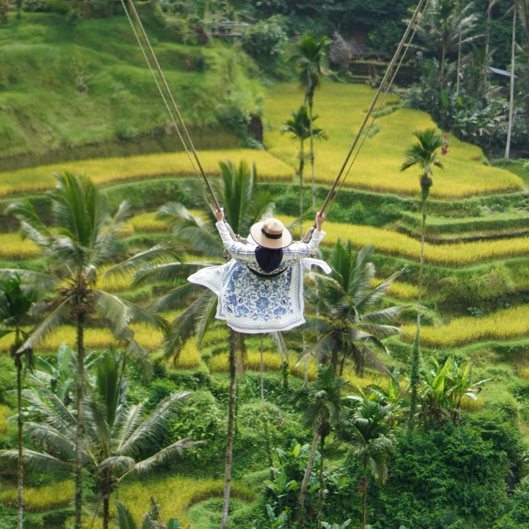 Traveller wearing a sun hat on a swing above green rice terraces and palm trees in Bali.